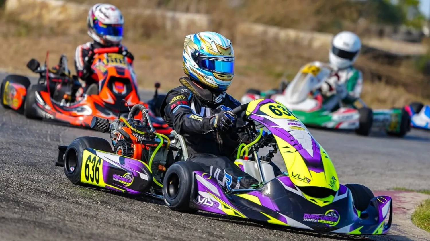 Kart racers competing on an outdoor track during a sprint race with the lead kart in purple and yellow livery